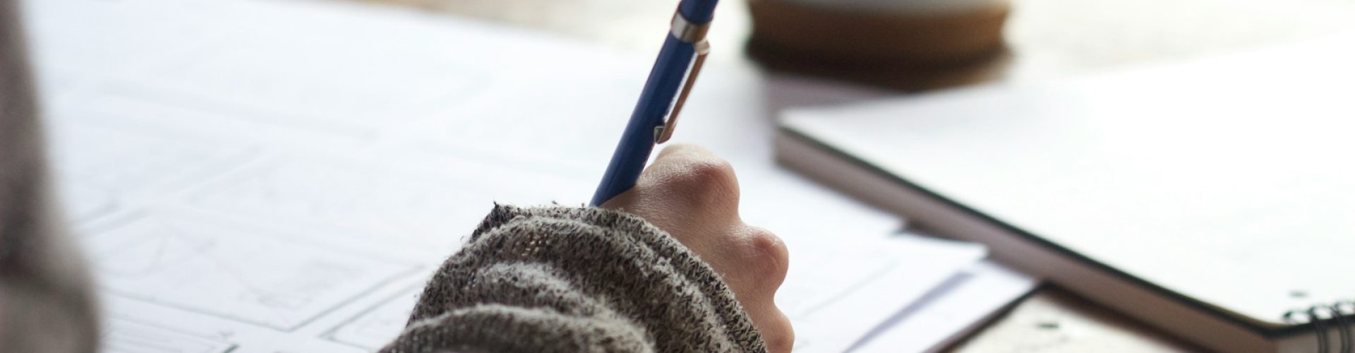person writing on brown wooden table near white ceramic mug